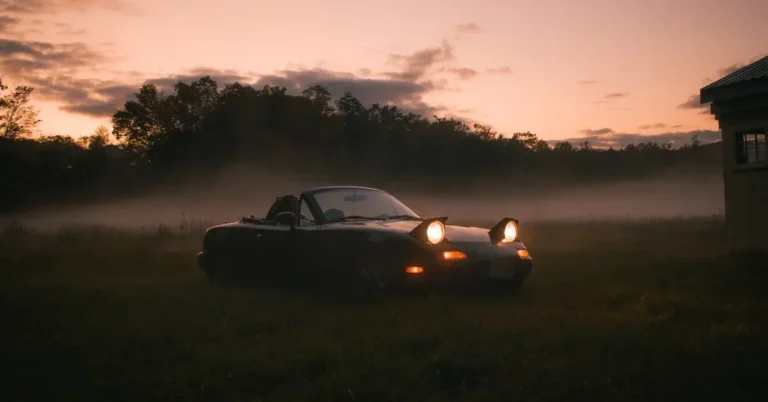 An Mazda Mx5 with open Headlights turned on standing on a meadow in front of the sunset