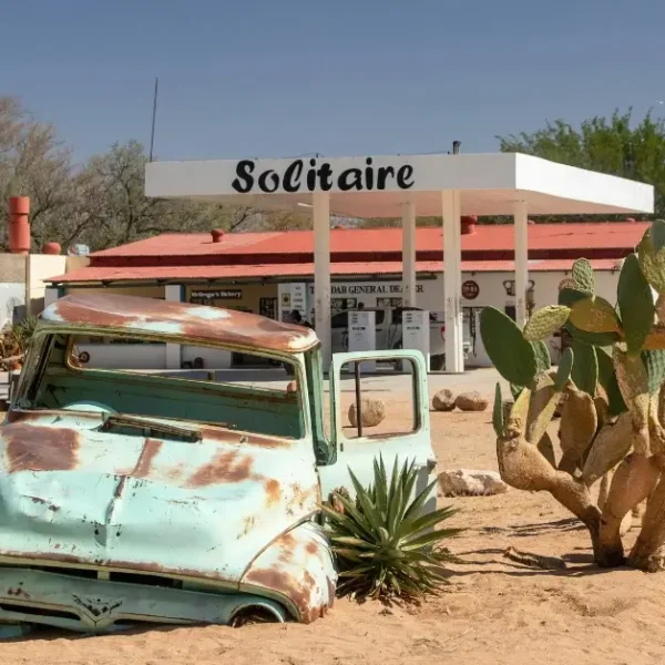 A rusted car without wheels in a desert like area at a gas station